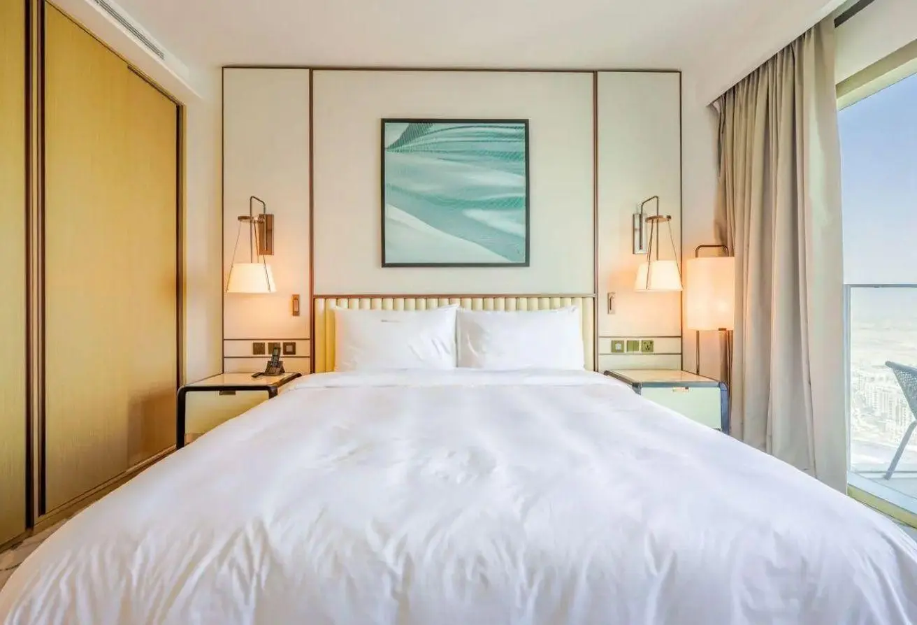 King-size bed in a modern hotel room with white linens, two bedside tables, and wall-mounted lamps; a framed abstract artwork above the headboard.