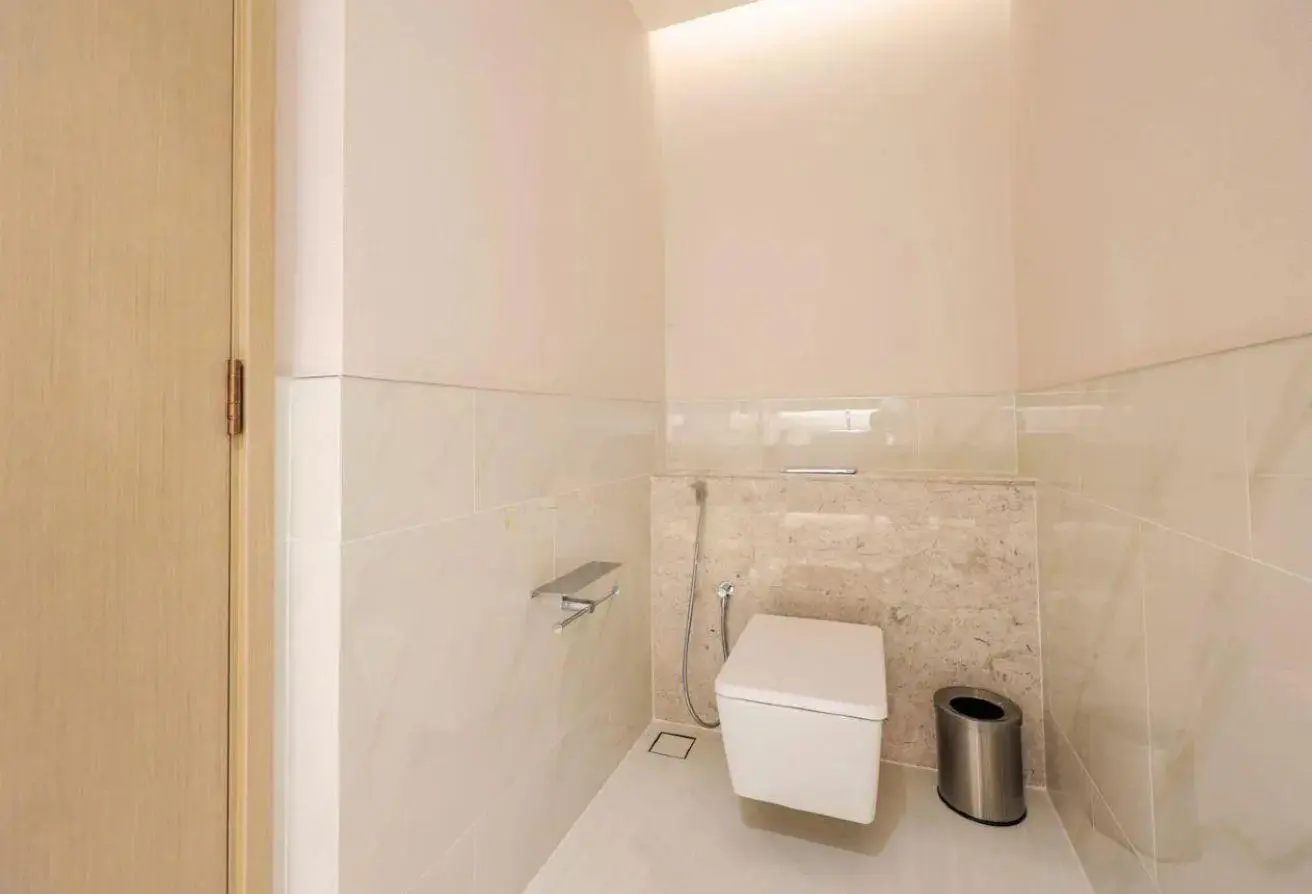 Minimalist bathroom: wall-mounted white toilet, handheld bidet spray, chrome toilet paper holder, and a silver trash can.