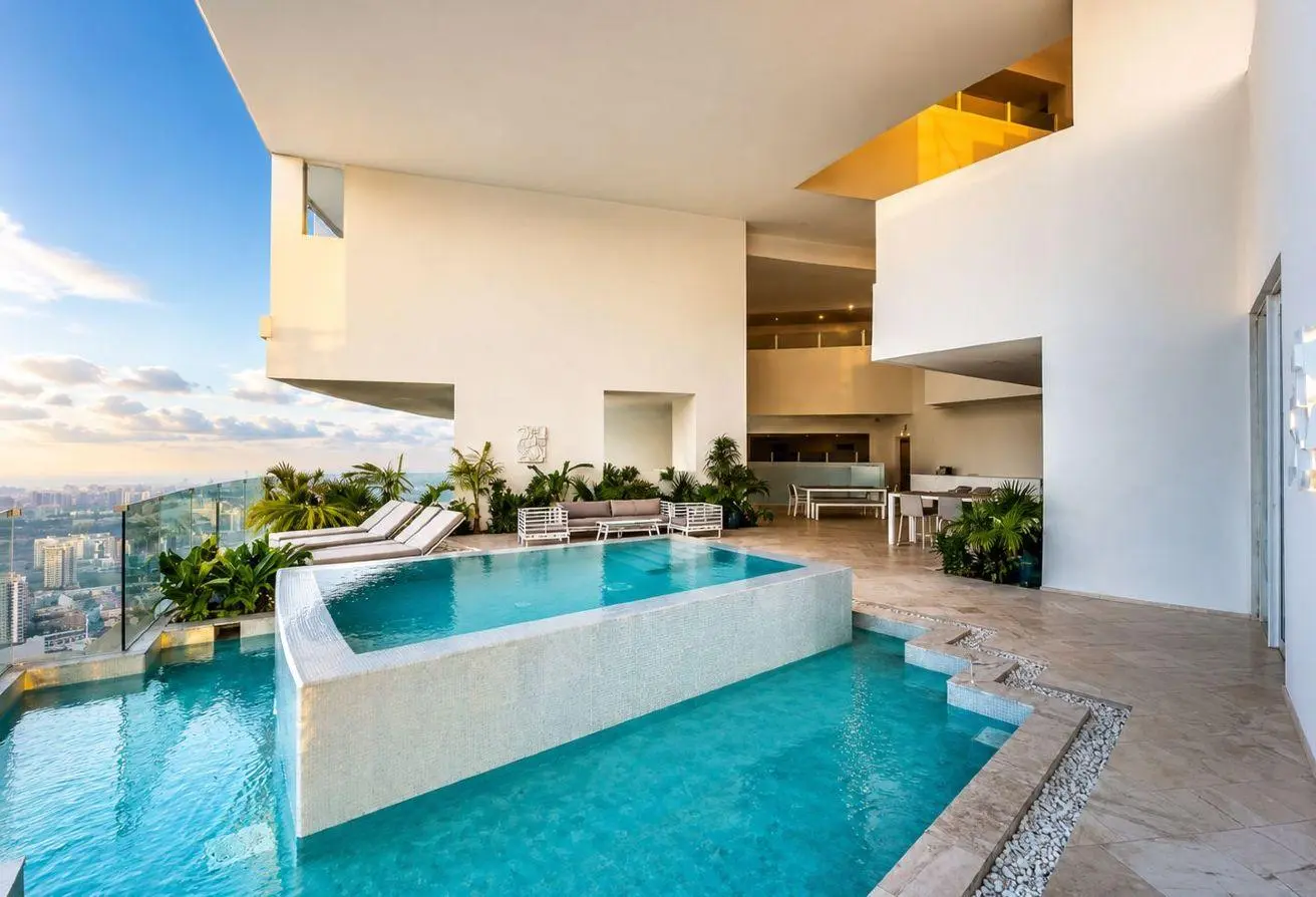 Rooftop pool area with bubbling turquoise water, lounge chairs, and glass railing overlooking a city skyline, modern white walls.