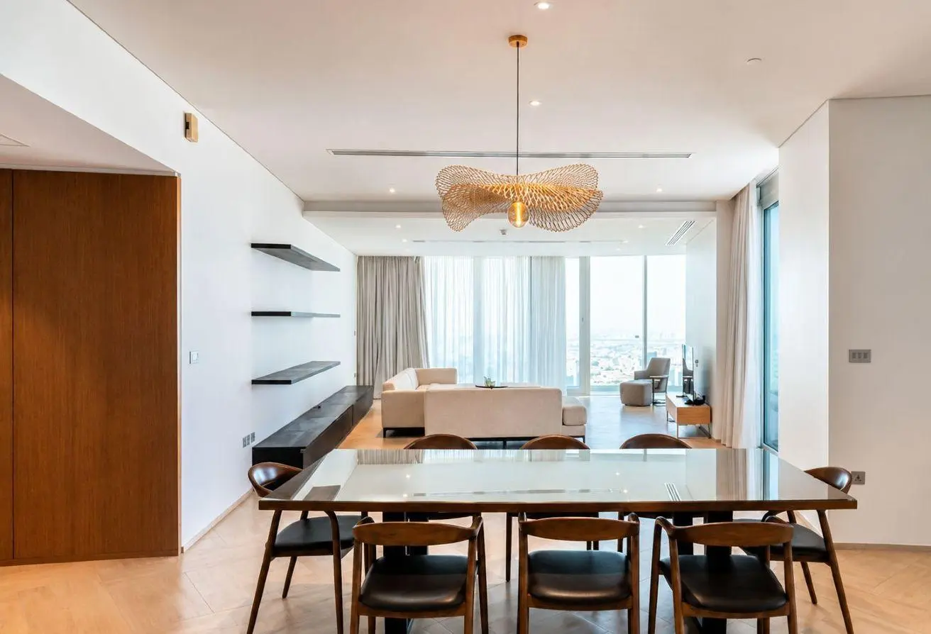 Open-concept dining and living area with a glass-top dining table and wooden chairs, beige sofa, and large windows with sheer curtains behind.