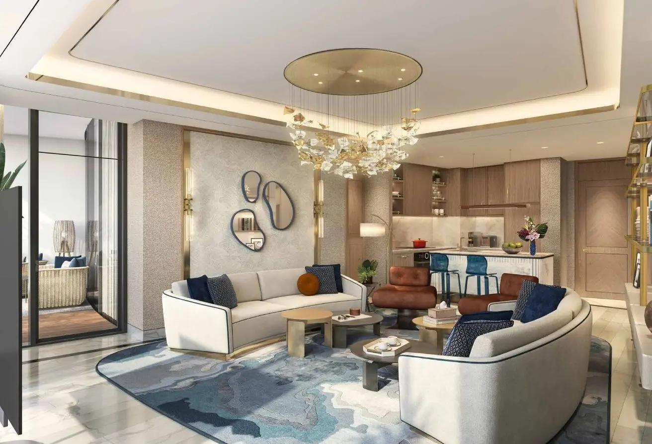 Modern open-concept living area with a curved white sofa, blue cushions, and round coffee tables; a blue-toned abstract rug anchors the space, and a decorative chandelier hangs from a recessed ceiling.Near the back, a contemporary kitchen with a marble island and blue bar stools is visible, connected to the living room through warm wood cabinetry and soft lighting.