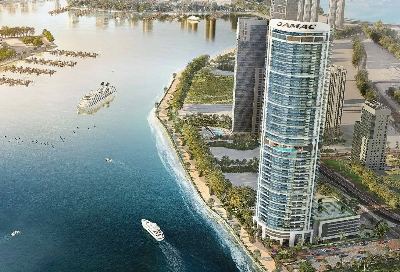 Tall curved glass skyscraper labeled DAMAC beside a waterfront promenade with boats and a coastline