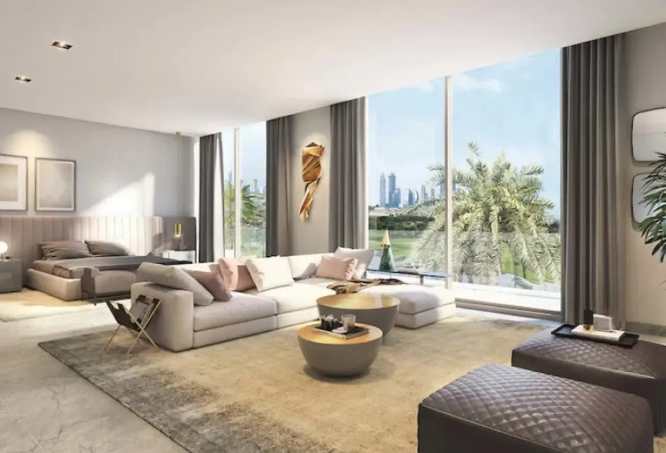 Bright modern living room with floor-to-ceiling windows showing a city skyline and palm trees, a light sofa with pink cushions, and two round coffee tables; bedroom area visible in the back.