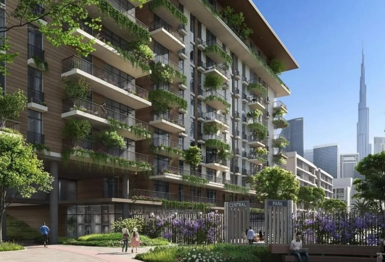 Modern apartment building with balconies full of greenery, a landscaped park area, and the city skyline in the background.