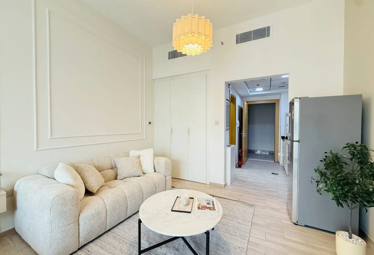 Bright minimalist living room with a beige sofa, round marble coffee table, and light wood floors; a large refrigerator is visible in the open kitchen area.