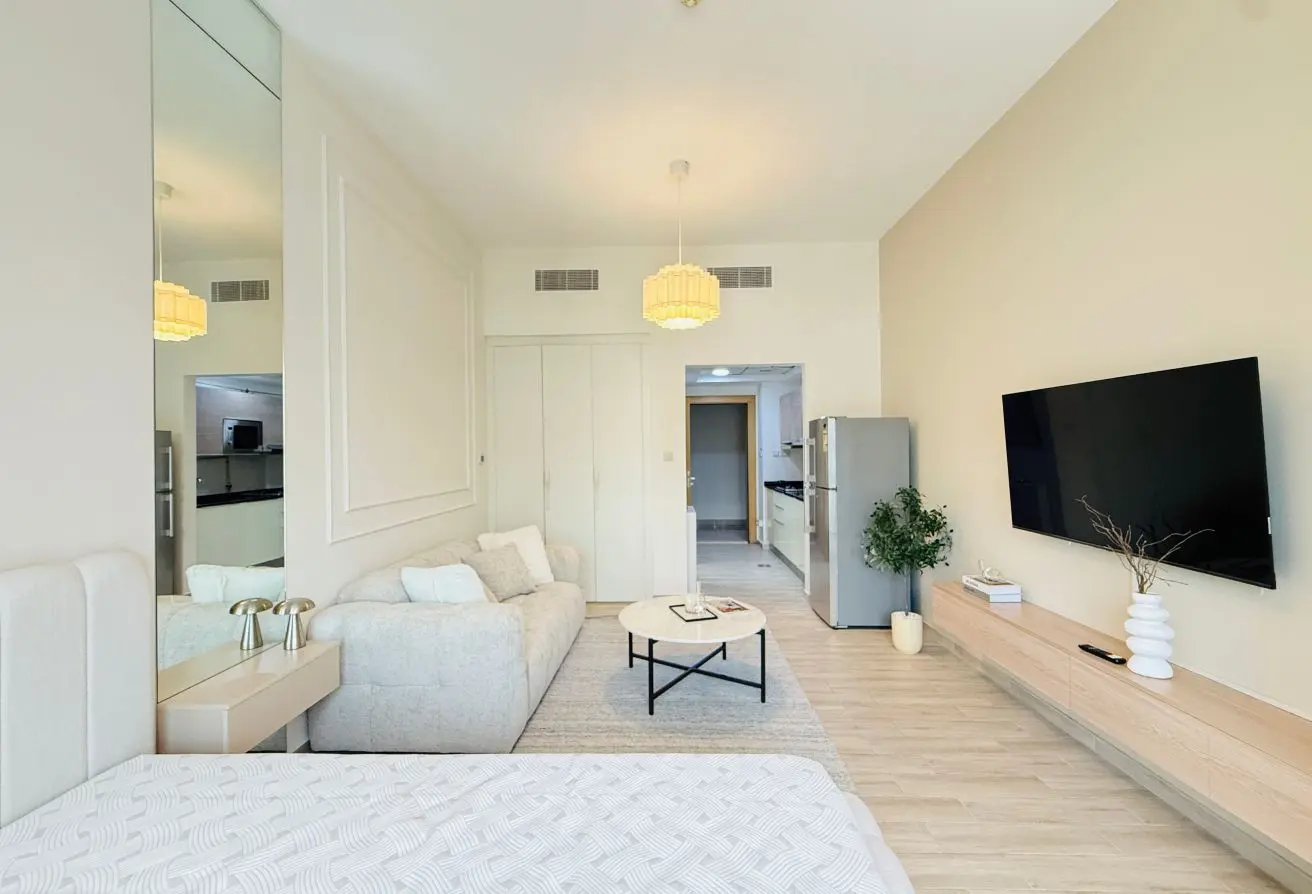 Bright open-plan living room with a wall-mounted TV, gray sofa, round coffee table, and a light wood console along the wall; kitchen visible in background.
