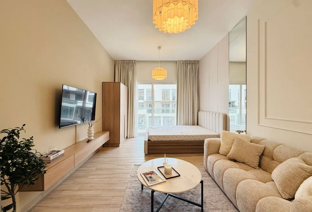 Bright modern studio living area with a bed in the background, beige sofa, round marble coffee table, and wall-mounted TV.