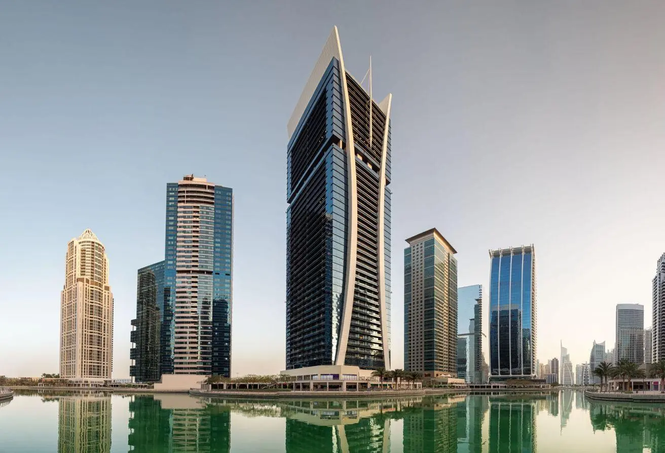 Skyscrapers along a calm waterfront with modern glass towers and their reflections in the water under a clear sky.