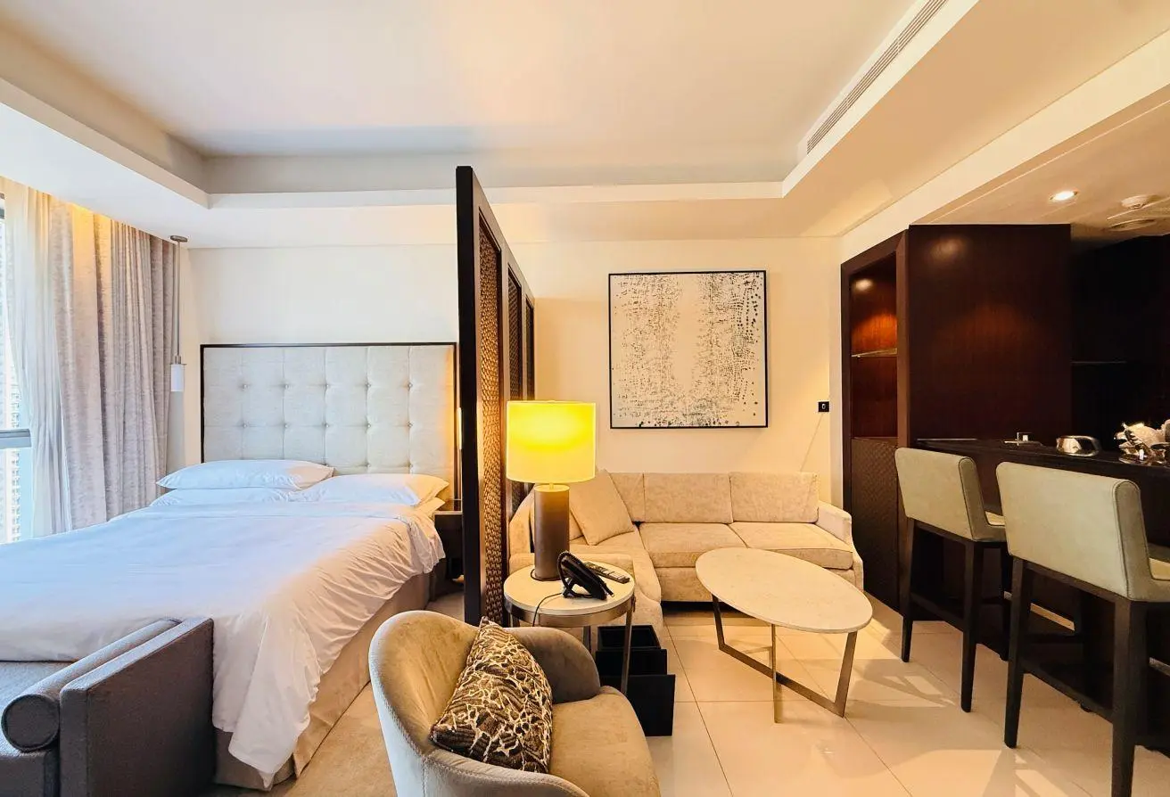 Hotel suite with a large bed, tufted headboard, and beige seating area nearby