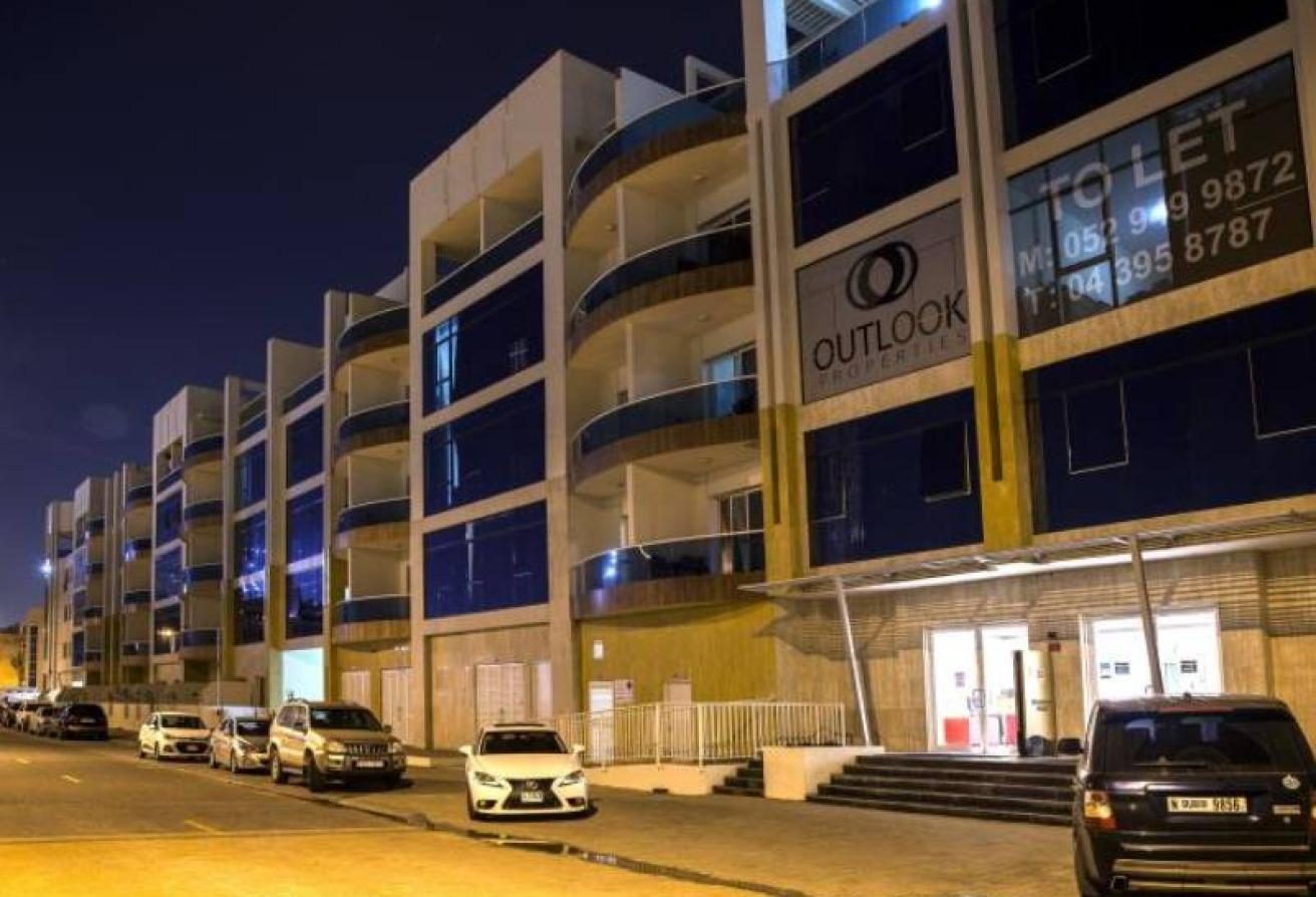 Nighttime street view of a modern apartment building with blue-tinted balconies and parked cars along the curb, signage reading 'OUTLOOK'.