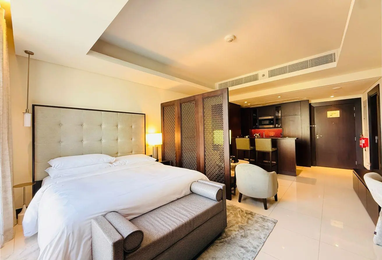 Hotel suite with a king-size bed, cushioned headboard, and seating area separated by a decorative partition.