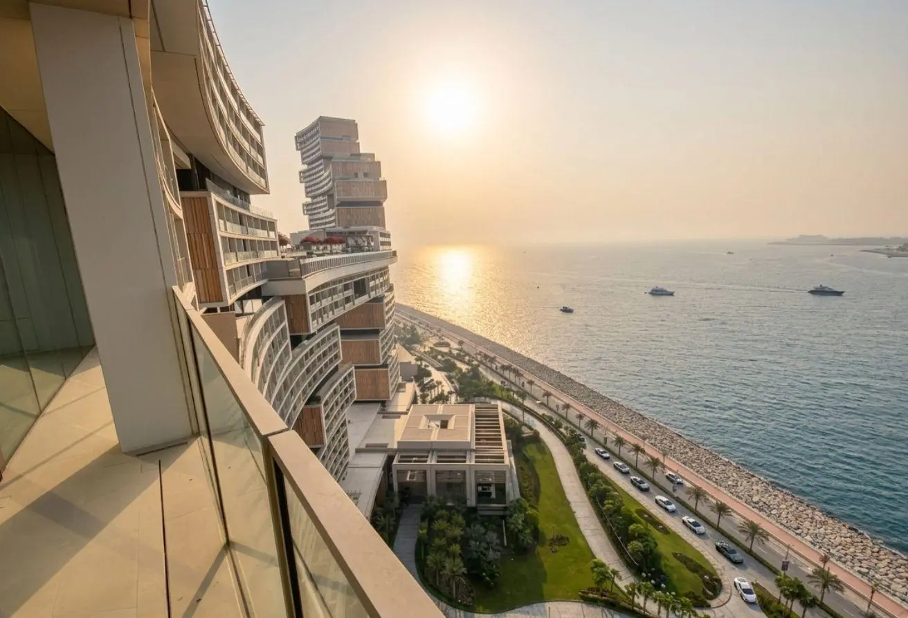 Sunset over a curved, glass-fronted hotel by the sea, with a palm-lined promenade and boats on calm water.