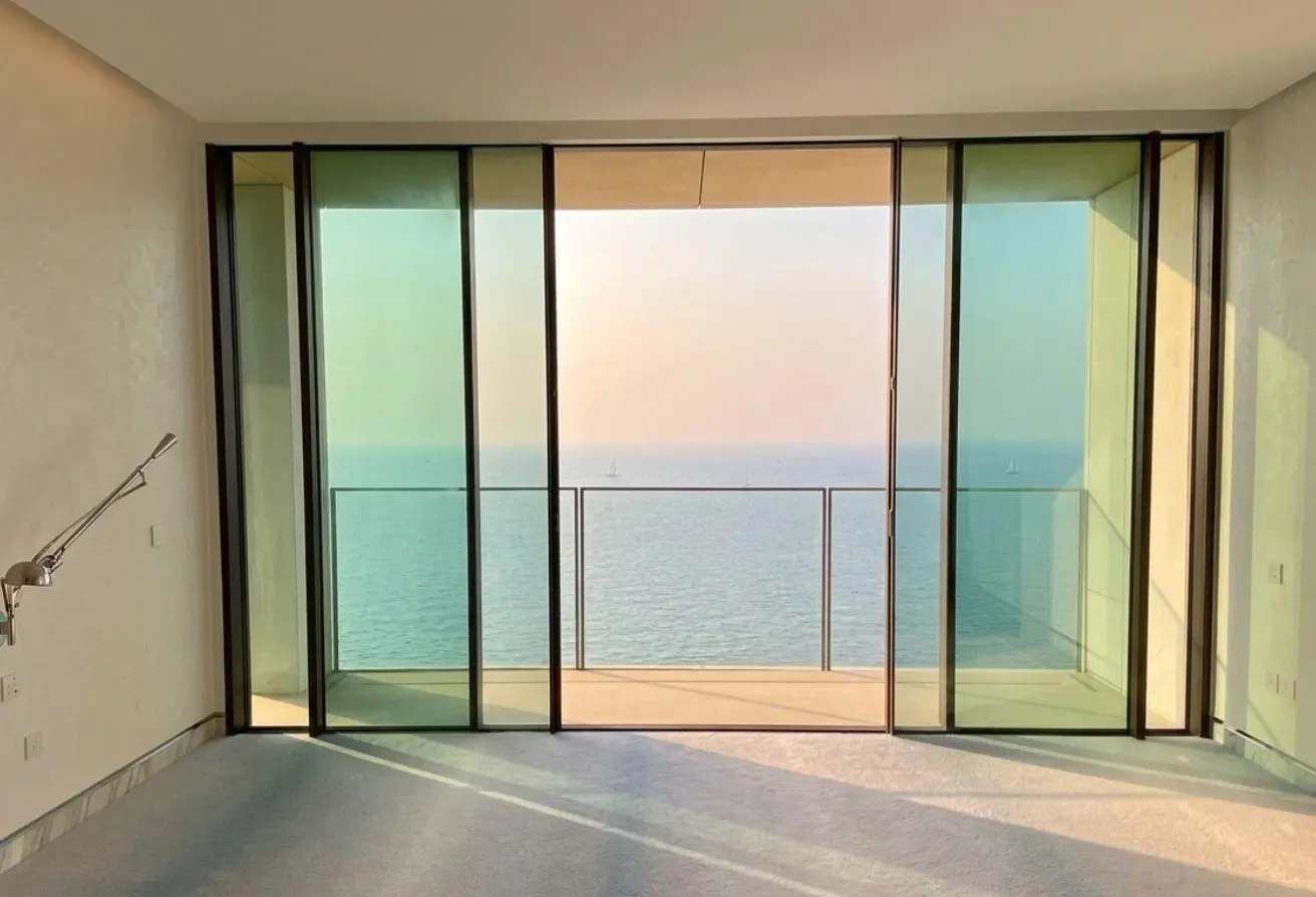 Bright empty room with large glass sliding doors opening to a balcony overlooking the ocean and a calm sea beyond the horizon at sunset.