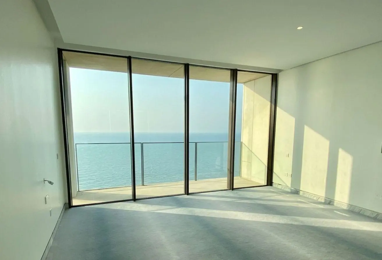 Empty, bright room with floor-to-ceiling glass doors opening to a balcony overlooking the ocean view at sea level.