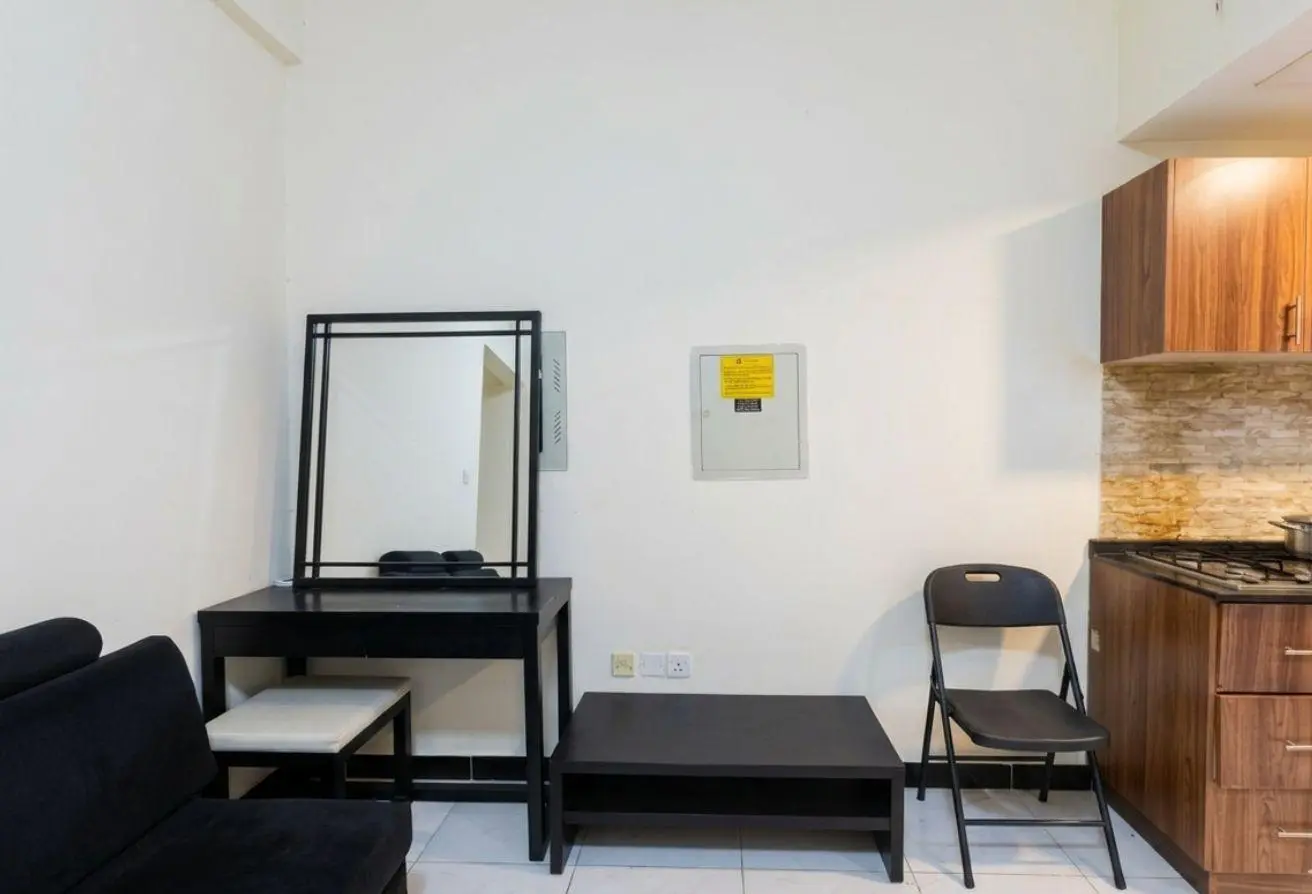 Small living area with black furniture: vanity table and mirror, bench, low coffee table, and a black folding chair near a kitchen with wood cabinets on the right.