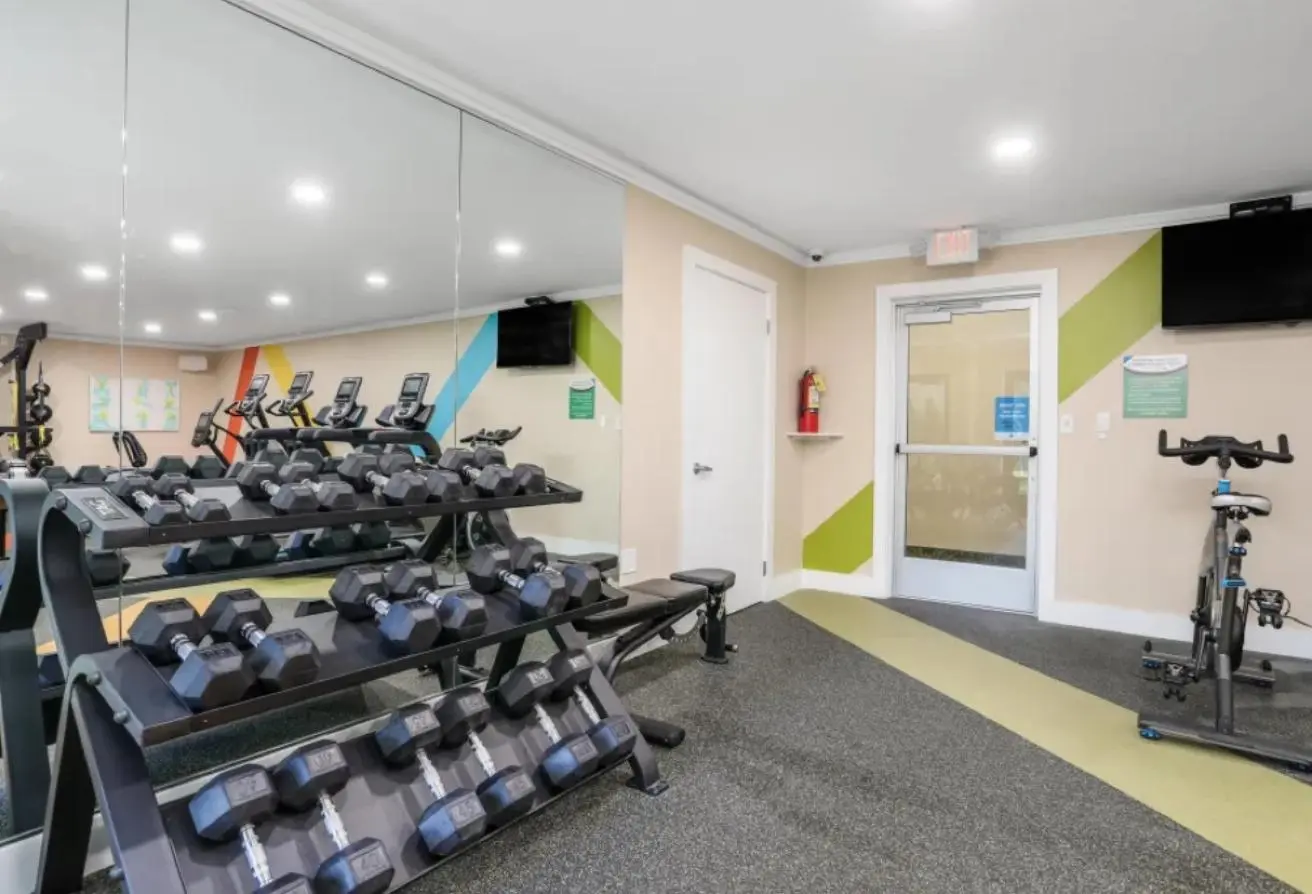 Small gym with a two-tiered rack of dumbbells in the foreground, cardio machines along a mirrored wall, and a stationary bike near a glass door with colorful wall accents and a fire extinguisher nearby.
