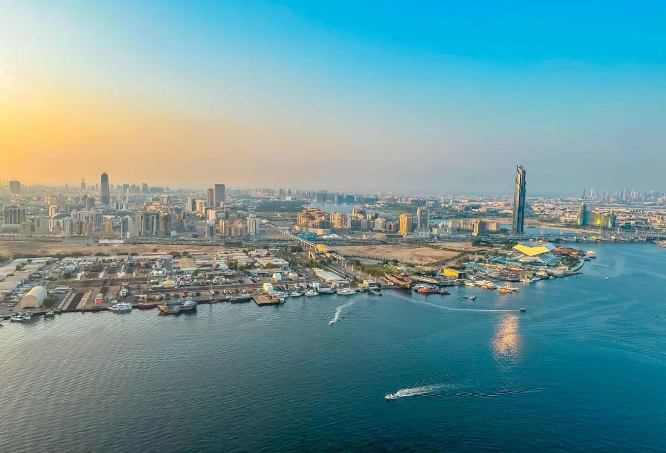 Aerial view of a coastal city at sunset with a skyline of high-rise buildings and a busy harbor with boats.