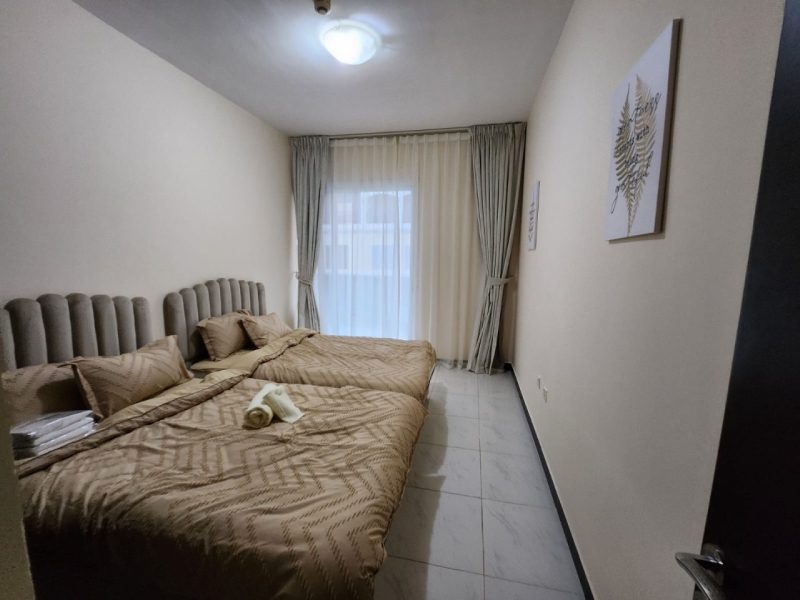 Twin hotel room with beige bedding, tufted headboards, and towels on one bed beneath a large window with sheer curtains.