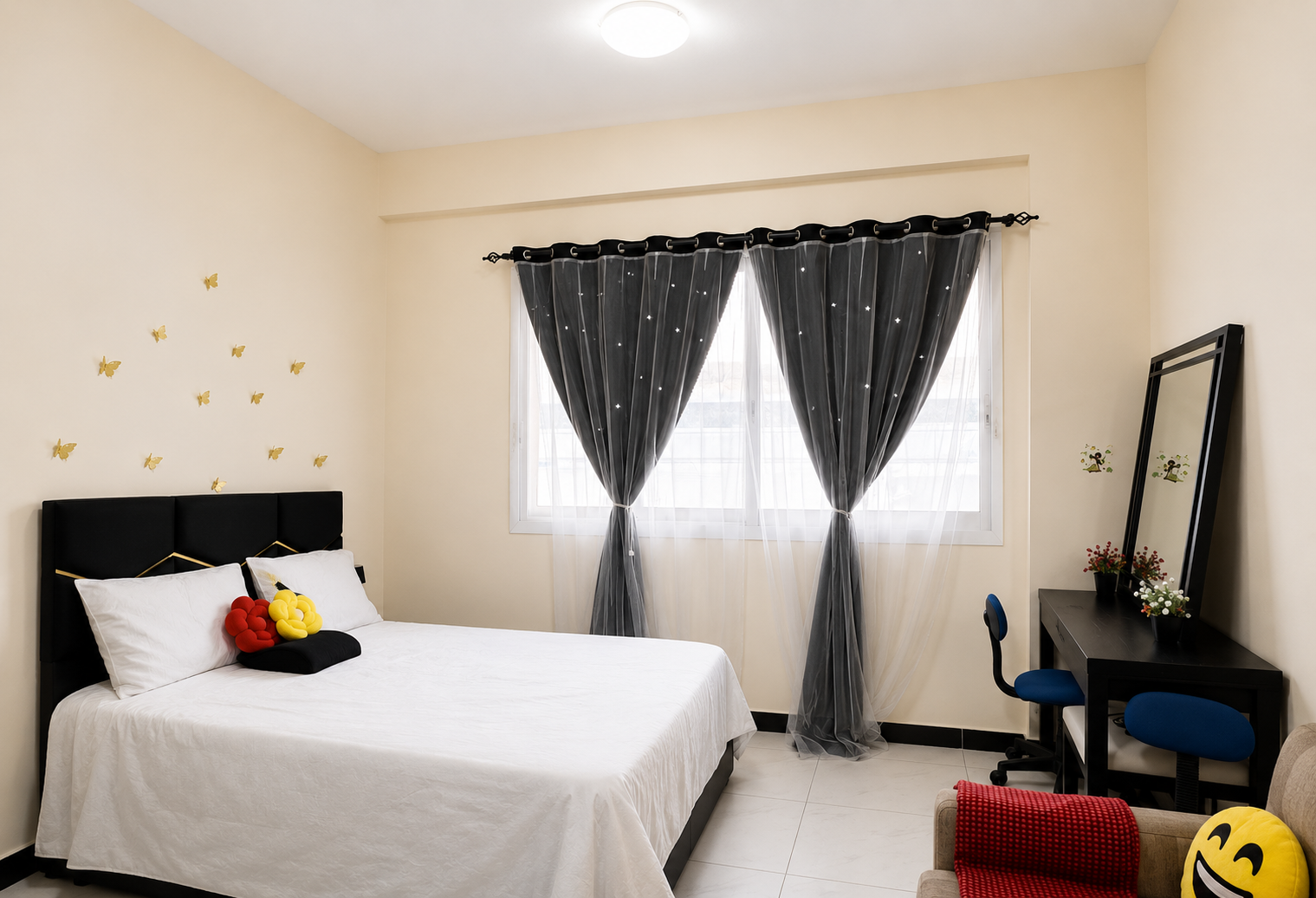 Bedroom with a white bed and black headboard, butterfly wall decals, and a window with sheer gray curtains; a desk and blue chair on the right.