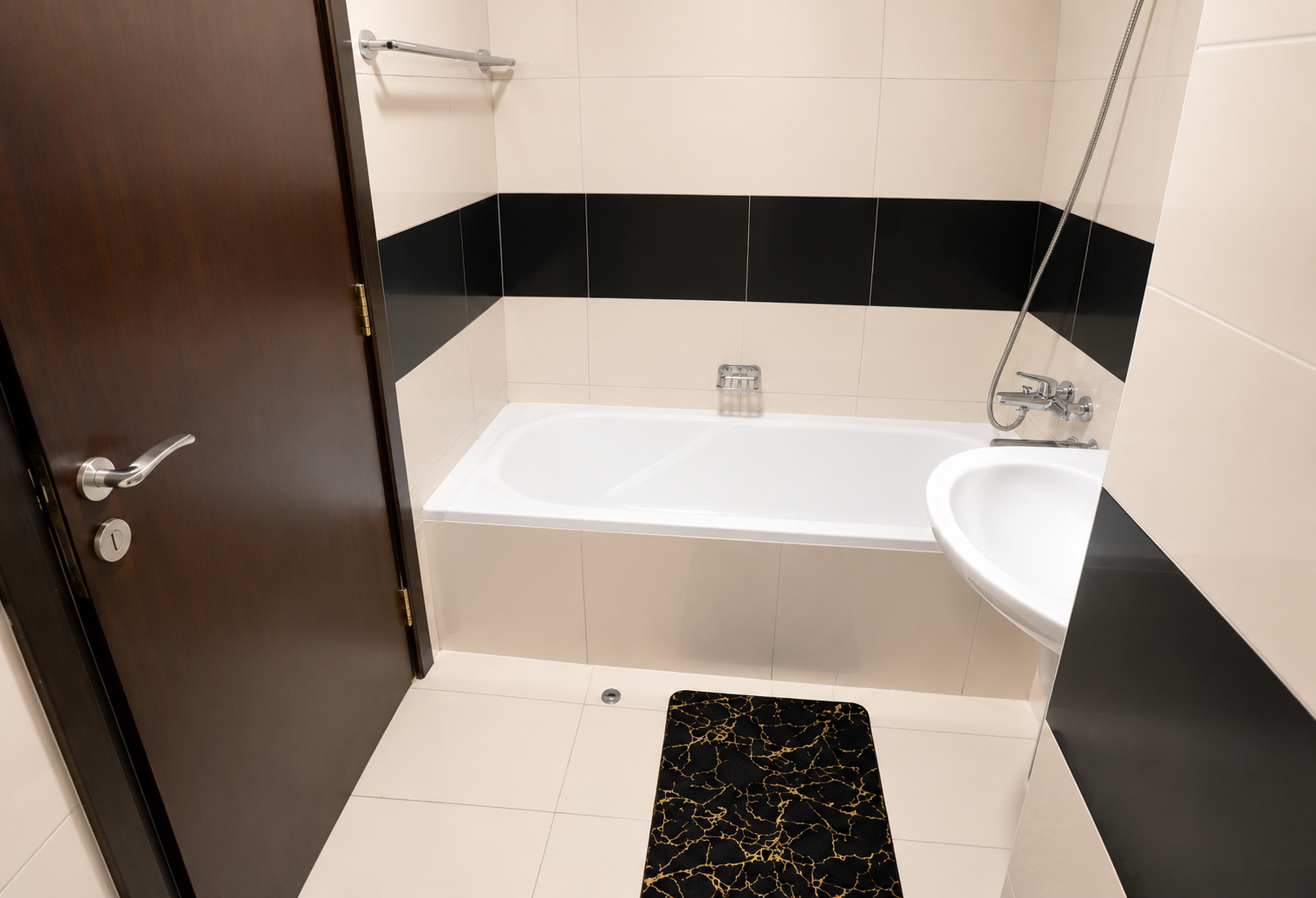 Modern bathroom with a white tub, white sink, beige tiles, and a dark wood door. A black horizontal tile stripe runs around the room above the tub and sink.