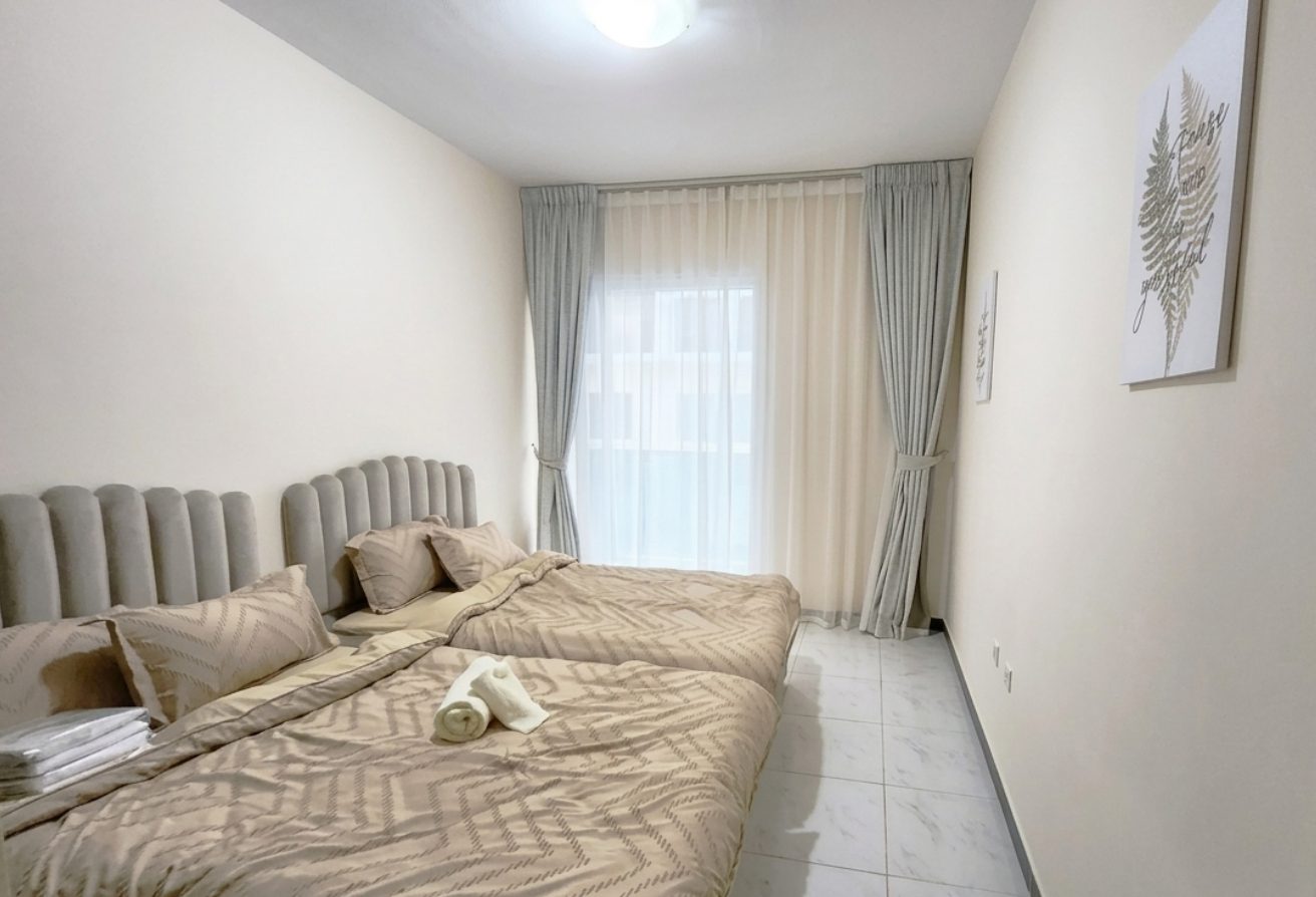 Twin bed hotel room with beige bedding, padded headboards, and sheer white curtains by a large window.