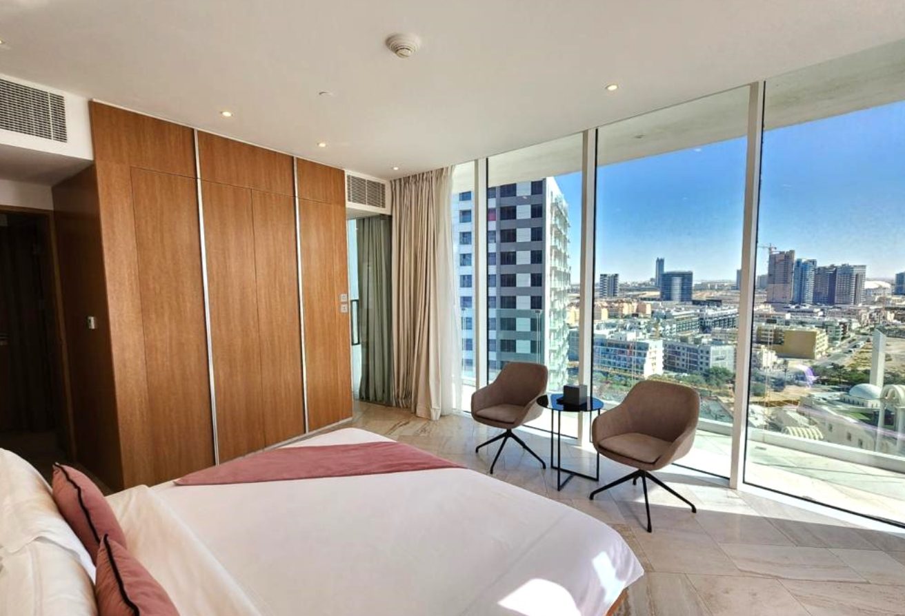Hotel bedroom with floor-to-ceiling windows showing the city skyline; two chairs and a small table by the window.