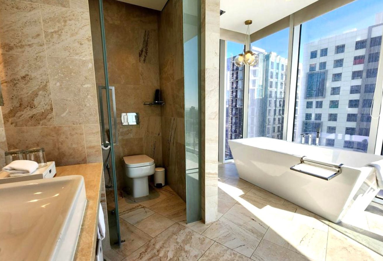 Luxurious bathroom with a white freestanding bathtub near floor-to-ceiling windows overlooking city buildings; bright daylight fills the room.