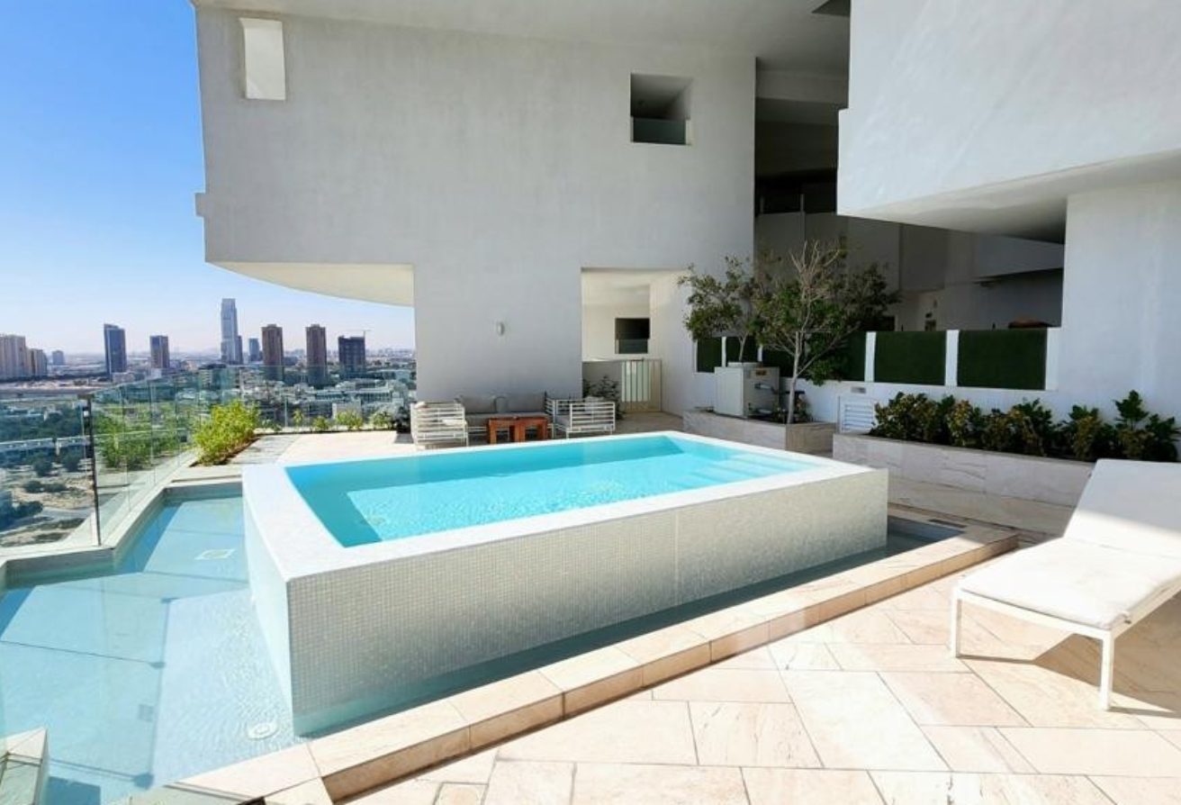 Rooftop pool on a white modern building with glass railing and city skyline in the distance; sunlit terrace with lounge chair.