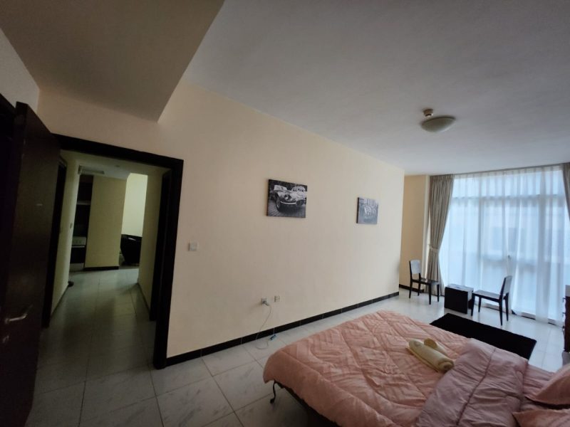 Hotel room with a pink quilted bed, a couple of wall art prints, and a large window with sheer curtains; small table and chairs nearby.