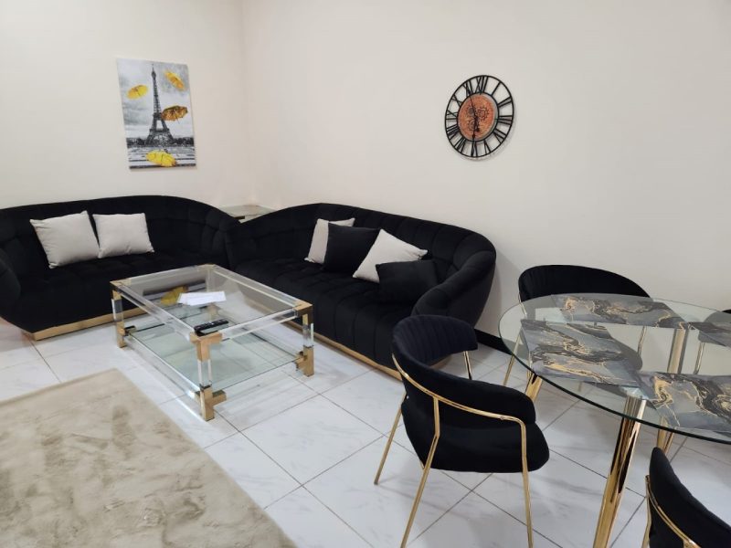 Modern living room with a black sectional sofa, glass coffee table, and a round marble dining table with black and gold chairs on white tile floors.