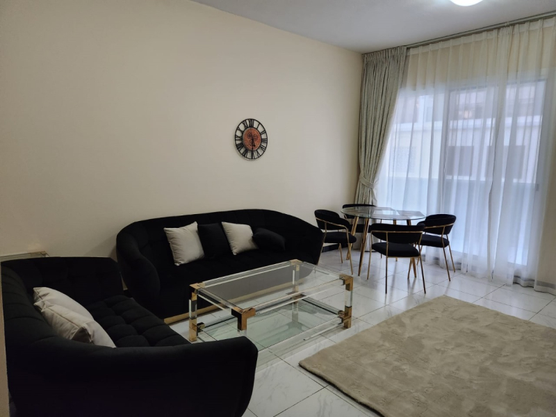 Modern living room with black curved sofas, glass coffee table, and a wall clock, adjacent to a dining table by large windows with sheer curtains.